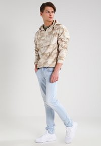 Pulóver de camuflaje beige con zip frontal y capucha ajustable, combinado con jeans de color azul claro y zapatillas deportivas blancas. Texturas suaves.