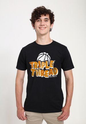 Jonge man met krullend haar die een zwart T-shirt draagt met de tekst "TRIPLE THREAD" en een basketbalafbeelding, staand tegen een effen witte achtergrond.