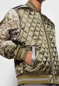 Homme portant une veste bomber matelassée en satin vert olive et beige avec des poignets côtelés rayés et une poche latérale, main partiellement dans la poche.