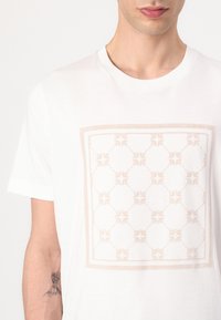 Camiseta de algodón blanca con un patrón gráfico decorativo de color beige claro enmarcado en un cuadrado, con una textura suave y cuello redondo.