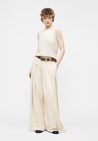 Άτομο που φοράει μπεζ αμάνικο τοπ και μπεζ παντελόνι με φαρδιά πόδια και καφέ ζώνη, στέκεται με το ένα χέρι στην τσέπη μπροστά σε απλό φόντο.