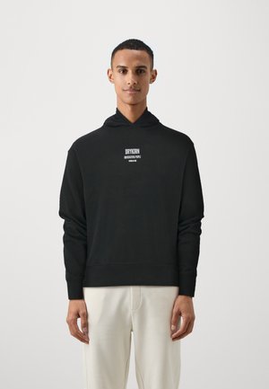 Black hoodie with a relaxed fit, featuring a front logo and ribbed cuffs. Fabric appears soft, worn with light-colored pants.