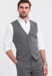 Gilet grigio texture con cinque bottoni, indossato sopra una camicia bianca. Presenta un design a V e tasche laterali per un aspetto su misura.