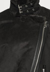 Zwarte faux-suède jas met een hoge kraag, contrasterende zwarte leren accenten en een diagonale ritssluiting. Gelaagde textuur en zilveren hardware.