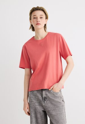 Jonge persoon met kort blond haar, gekleed in een koraalroze T-shirt en grijs gewassen jeans, poseert met één hand in de zak tegen een witte achtergrond.