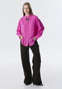 Camisa de botón de color rosa brillante con un bolsillo delantero, combinada con pantalones de pierna ancha de estampado de leopardo marrón. Detalle de collar con una cadena negra adicional.