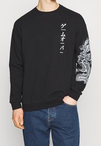 Černá mikina s dlouhými rukávy s bílým japonským textem na přední straně a podrobným bílým obrázkem draka na pravém rukávu.