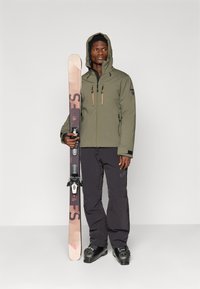 Lyžiarska výbava zahŕňajúca vodoodolnú bundu olivovej farby so zipsovými vreckami a čierne lyžiarske nohavice. Lyžiar drží ružovú a čiernu lyžu s viazaním.