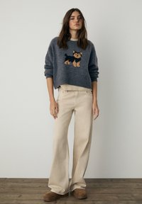 Pull gris en maille avec un motif de chien, associé à un jean beige à jambes larges et des chaussures marron texturées. Le mannequin pose devant un fond uni.