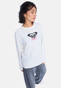 Biała koszula z długim rękawem z logo czarnego serca i napisem "ROXY" w kolorze różowym. Modelka ma na sobie szare, luźne dżinsy, prezentując swobodny wygląd.