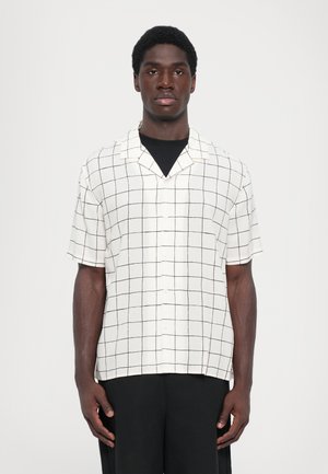 Korte mouw, geknoopte overhemd van witte stof met zwart rasterpatroon. Voorzien van een knoopsluiting aan de voorkant en een ontspannen pasvorm. Gekoeld met zwarte broek.