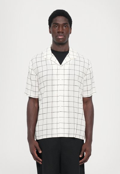 Chemise à manches courtes, col polo en tissu blanc avec un motif à carreaux noirs. Dotée d'un bouton sur le devant et d'une coupe décontractée. Portée avec un pantalon noir.