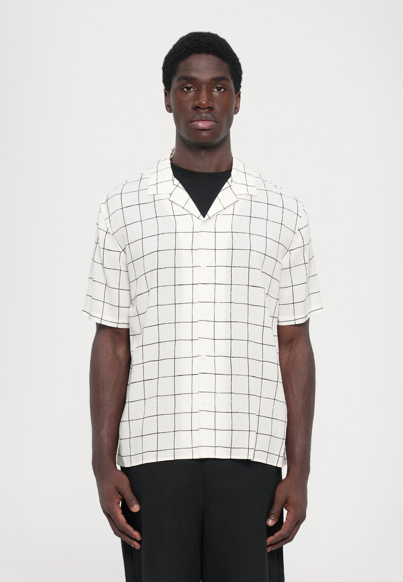 Korte mouw, geknoopte overhemd van witte stof met zwart rasterpatroon. Voorzien van een knoopsluiting aan de voorkant en een ontspannen pasvorm. Gekoeld met zwarte broek.