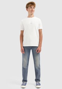 Tienerjongen die een wit T-shirt met korte mouwen met kleine tekst, blauwe jeans en blauwe sneakers draagt, staand tegen een effen lichte achtergrond.