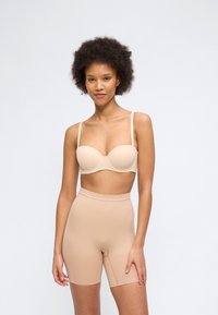 Γυναίκα με σγουρά μαλλιά που φοράει μπεζ στράπλες σουτιέν και μπεζ σορτς σμιλεύσης με ψηλή μέση, σε απλό φωτεινό φόντο.