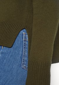 Maglione lavorato a maglia verde oliva con polsini a coste, spacchi laterali e texture morbida, abbinato a jeans blu. Dettaglio tasca visibile sui jeans.