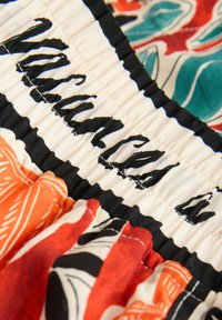 Elastický pás s čiernym prešitým názvom značky na bielom materiáli, pripojený ku farebnému kvetinovému materiálu v odtieňoch oranžovej, červenej, tyrkysovej a čiernej.