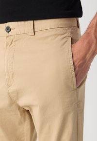 Beige broek van lichtgewicht stof, met een knoopsluiting, zijzak en een gladde textuur met minimale stikseldetails.