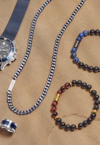 Silver klocka med blå mesharmband, blå och svarta pärlarmband, silver- och blå ring, samt ett silverhalsband på en beige yta.