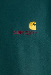 Tamnozelena tkanina s izvezenim žutim 'C' logom i 'carhartt' u burgundiju, s teksturiranim završetkom.