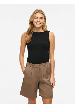 Ženska se smeji, oblečena v črno brezrokavnik in ohlapne rjave kratke hlače z visokim pasom, stoji z rokami v žepih pred enobarvnim ozadjem.