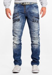 Jean en denim bleu avec plusieurs poches zippées, des zones décolorées et des détails cousus. Associés à des baskets blanches.
