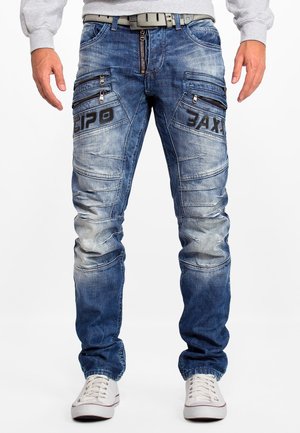 Jean en denim bleu avec plusieurs poches zippées, des zones décolorées et des détails cousus. Associés à des baskets blanches.