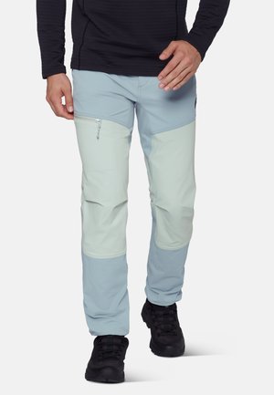 Mężczyzna w jasno-niebiesko-kremowych spodniach z podziałem kolorów, z zapinaną na zamek kieszenią na udzie, i czarnych butach turystycznych, idący do przodu.