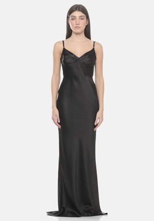 Donna con capelli castani lisci che indossa un lungo vestito di raso nero con spalline sottili, in piedi davanti a uno sfondo bianco semplice.