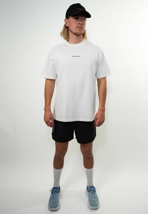 Homme aux cheveux blonds mi-longs, portant une casquette noire, un t-shirt blanc "Toujours Actif", un short noir, des chaussettes blanches et des baskets bleues, debout.