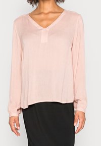 Blusa rosa claro de manga larga con escote en V y detalle de pliegues finos. Hecha de una tela texturizada, la parte inferior cae a la altura de la cintura, combinada con una falda negra.