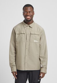 Beige overhemd met lange mouwen, voorzien van twee voorzakken en een knoopsluiting. Gemaakt van textuurstof met een logo op de linkerkant van de borst.