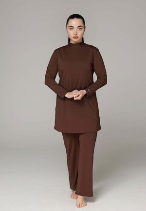 Femme debout pieds nus portant une tunique marron à manches longues et un pantalon large assorti sur un fond uni gris clair.