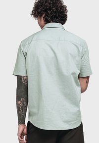 Hellgrünes Kurzarmhemd mit Kragen, glatter Textur und Rückenteil mit Schulterpassen. Stylisch mit geradem Saum, getragen mit dunklen Hosen.