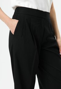 Zwarte broek met hoge taille, plooien en zijzakken. De stof heeft een gladde textuur en zorgt voor een op maat gemaakte en gestructureerde uitstraling.