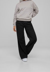 Lichtgrijze hoodie met een voorzak, gecombineerd met loszittende zwarte broek. Witte sneakers maken de outfit compleet. Effen achtergrond.