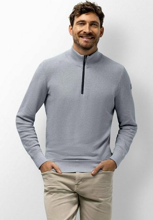 Man smiling with hands in pockets wearing light gray quarter-zip sweater and beige pants against a plain background.