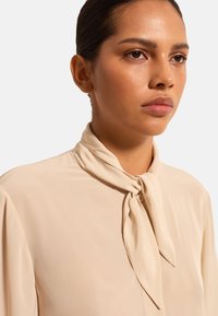Beige Bluse aus glattem Stoff mit gebundenem Schal-Kragen und Knopfverschluss, mit sanften Falten an den Schultern.