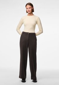 Kobieta stojąca z rękami za plecami, ubrana w kremowy prążkowany top z długim rękawem, ciemne szerokie spodnie i czarne buty na białym tle.