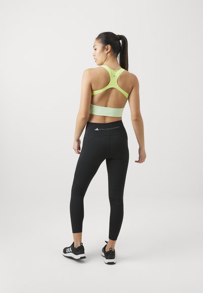 Reggiseno sportivo verde chiaro con una fascia larga e design a schiena scoperta; abbinato a leggings neri a vita alta con logo e dettagli perforati.