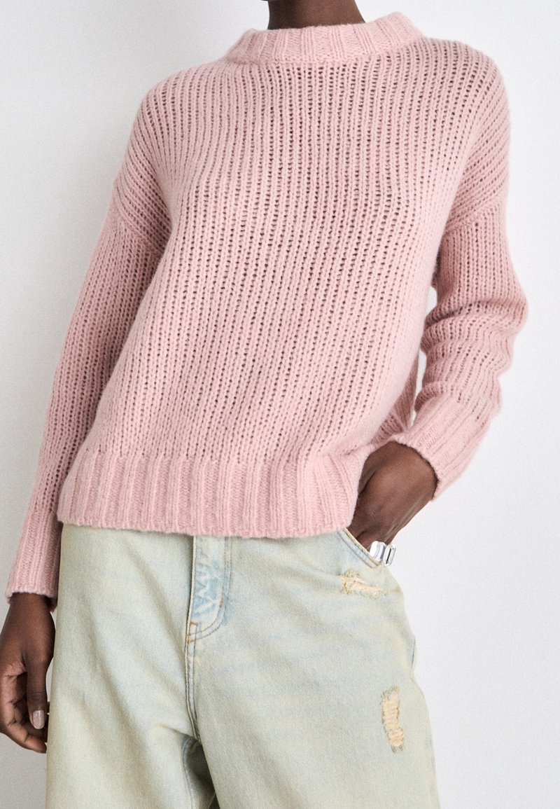 Personne portant un pull épais tricoté rose clair à manches longues et un jean délavé clair usé, main dans la poche sur fond uni.
