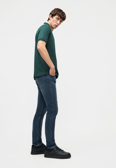Νεαρός άνδρας όρθιος πλαγίως με τα χέρια στις τσέπες, φοράει σκούρο πράσινο πόλο μπλουζάκι, μπλε τζιν και μαύρα αθλητικά παπούτσια σε ουδέτερο φόντο.