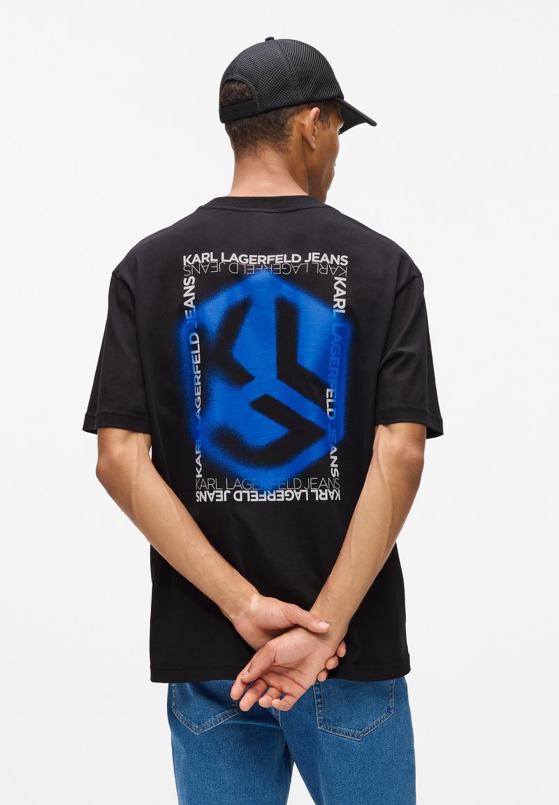 Melna T-krekls ar zilu grafisko logotipu aizmugurē, ko ieskauj balta teksts. Modelis valkā zilas džinsus un melnu cepuri.