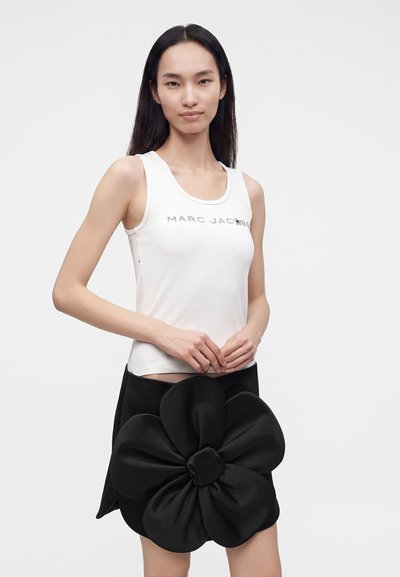 Femme portant un haut blanc sans manches avec le texte "Marc Jacobs" et une jupe noire ornée d'une grande fleur en tissu à l'avant.