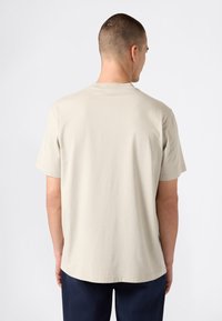Hombre vestido con una camiseta de manga corta beige y pantalones oscuros, de pie de espaldas contra un fondo blanco liso.