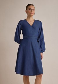 Denim omslagskjole i dyp blå med lange ermer, v-hals, belte i midjen og utsvingt skjørt. Myk tekstur, glatt finish og minimal detaljering.