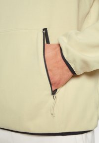 Hand placerad i den zip-försedda fickan på en beige fleecejacka med svart kant runt dragkedjan och ärmsluten.