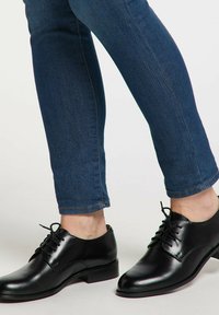 Person iført mørkeblå skinny jeans og polerede sorte læder snørebukser på en lys baggrund.