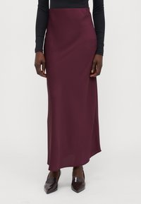 Gonna maxi in satin bordeaux con una texture liscia e uno spacco laterale, abbinata a scarpe lucide e nere a punta, indossata con un top nero aderente.