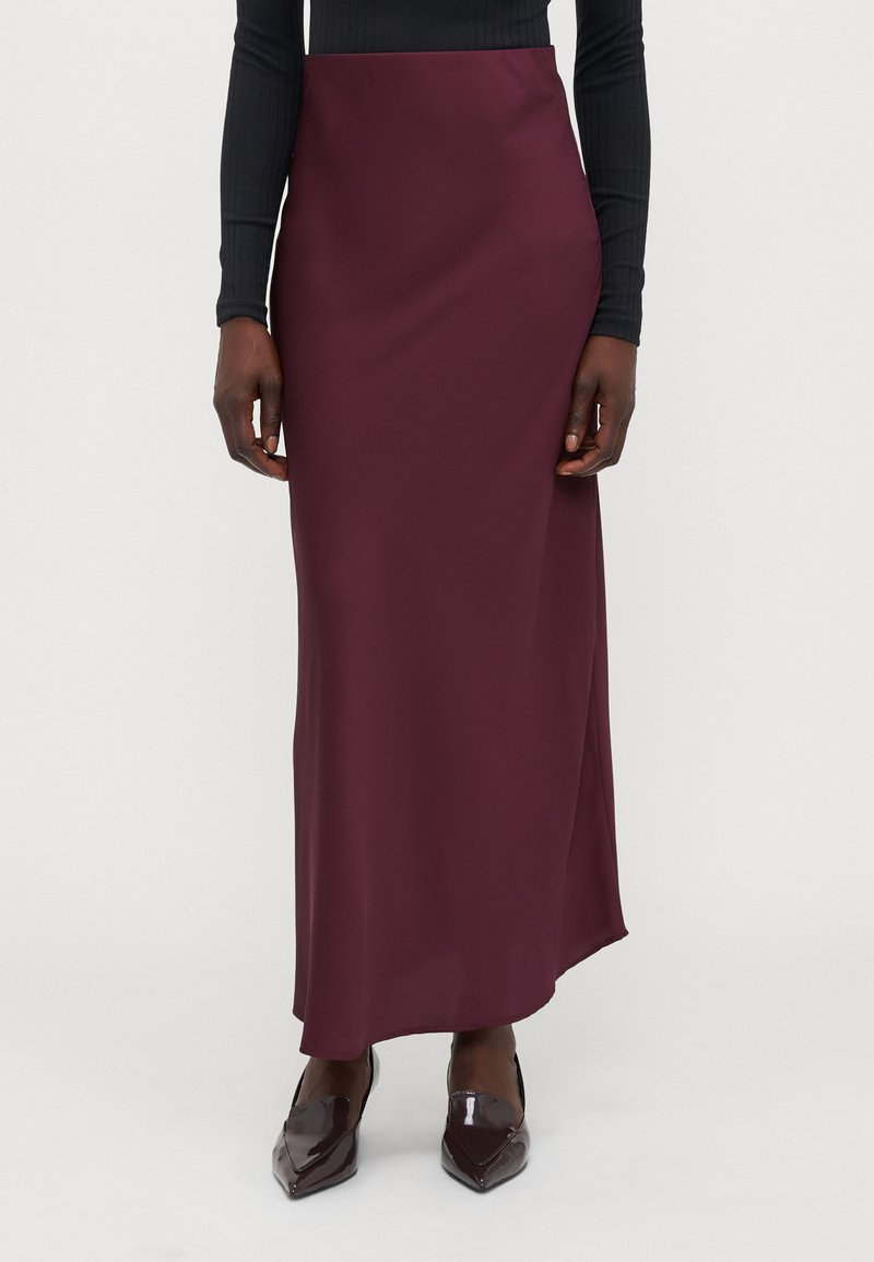 Gonna maxi in satin bordeaux con una texture liscia e uno spacco laterale, abbinata a scarpe lucide e nere a punta, indossata con un top nero aderente.